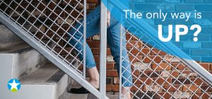 Untere Hälfte einer Dame in Jeans und schwarzen flachen Schuhen sichtbar: Sie geht eine Beton Treppe hinauf. Davor ein Stahlgeländer.